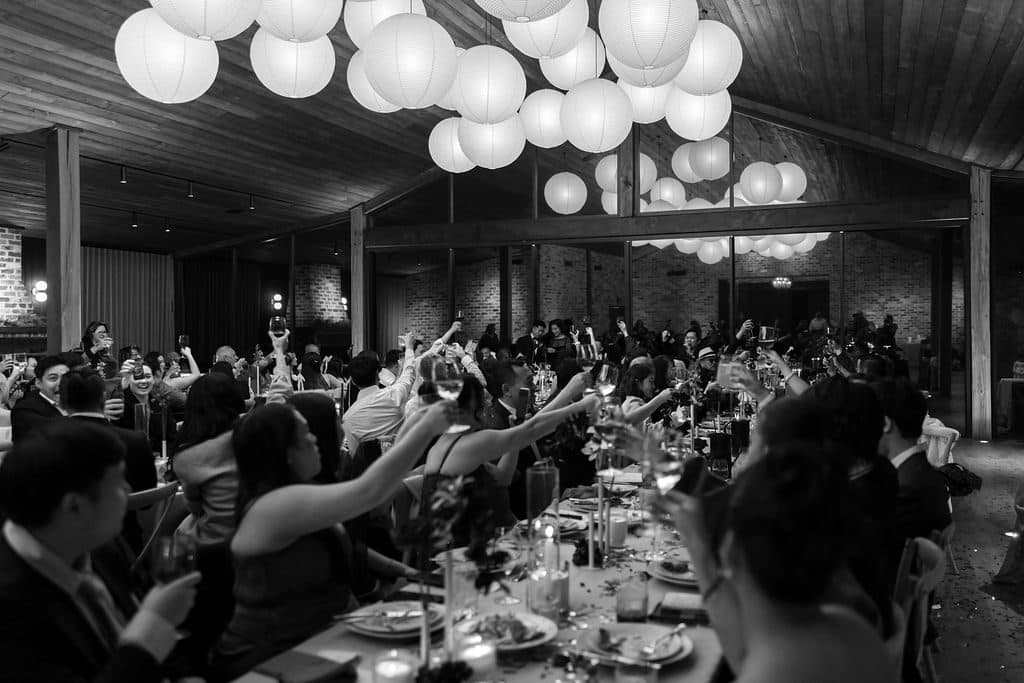 Guests on their feet at the long tables under the paper lanterns, in black and white