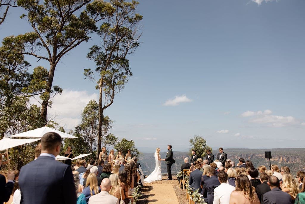 The ceremony seen from the back of the aisle: guests on timber benches either side, the couple holding hands at the cliff edge, and the escarpment stretching away behind them