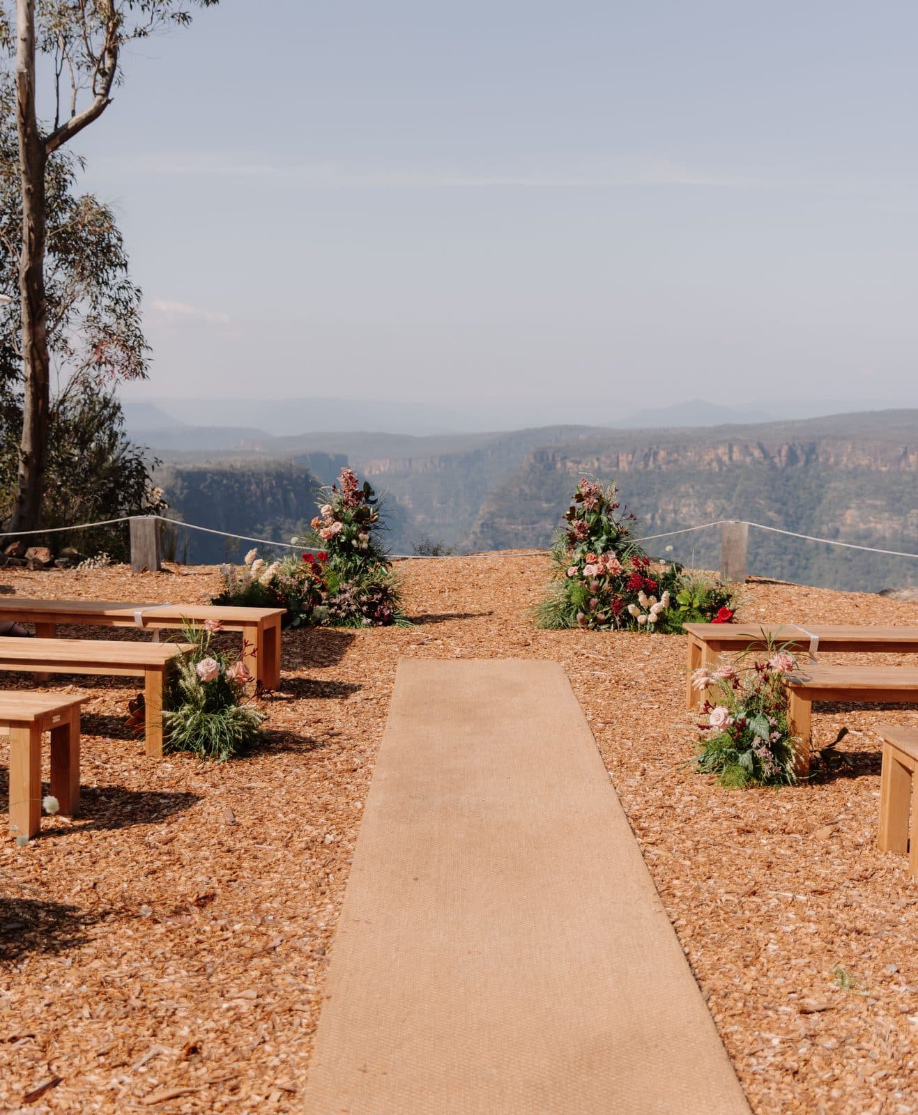 The clifftop ceremony site set for a wedding: timber benches either side of a jute runner, florals at the top of the aisle and the rope line, with the Shoalhaven gorge beyond