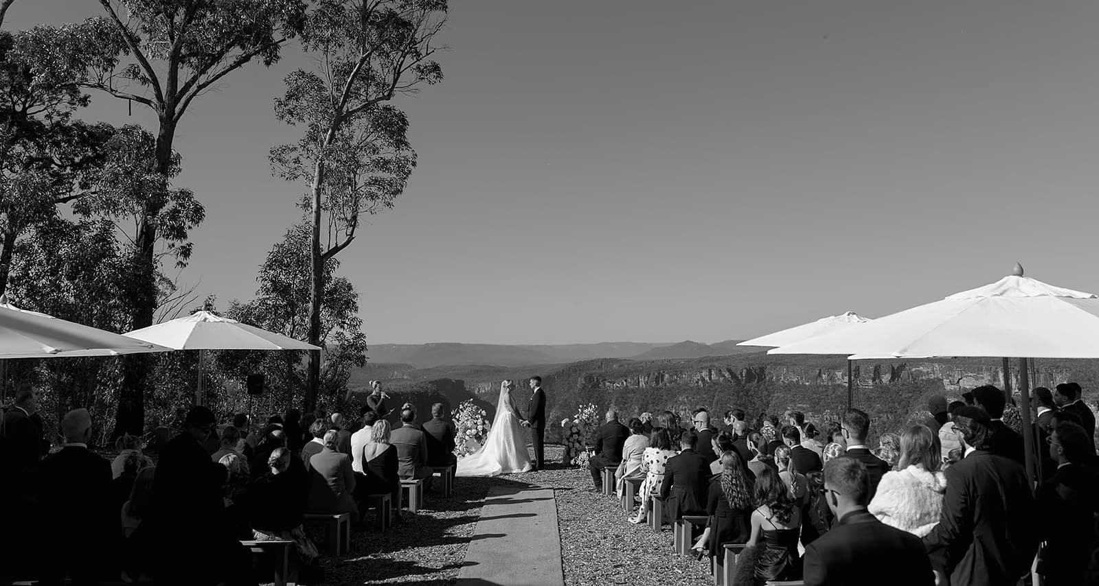 A ceremony on the open ledge at The Cliffs, guests seated on timber benches and standing under umbrellas, the Shoalhaven gorge falling away behind