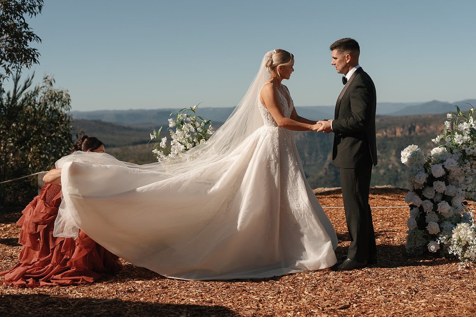 A couple holding hands at the cliff edge before their ceremony, white florals either side and the gorge stretching away behind them