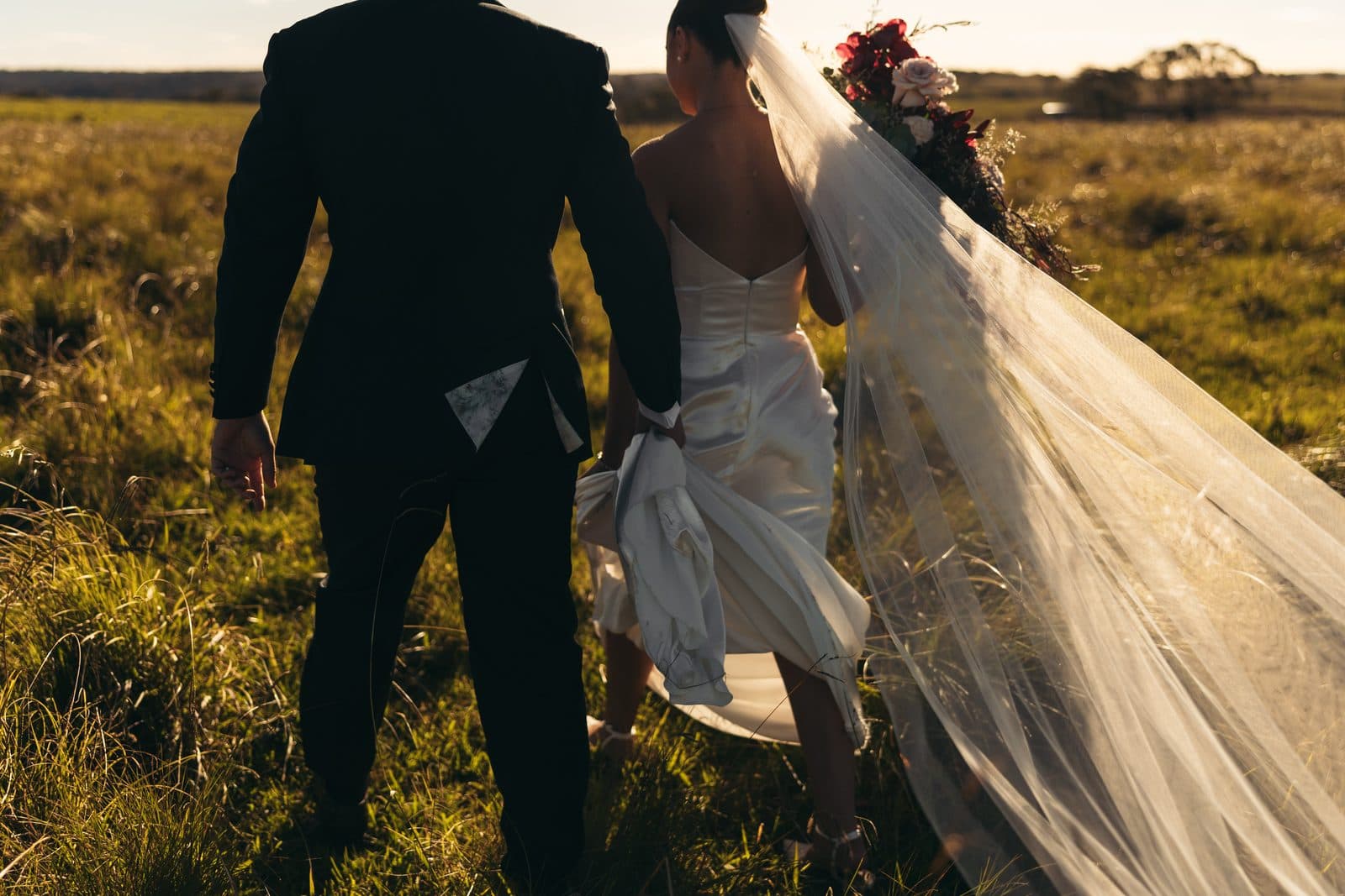 Ryan and Liv walking away through the long grass in the late afternoon, her veil trailing