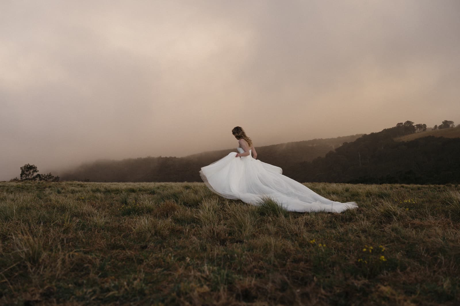 A model in a full tulle Moira Hughes gown standing in an open paddock at Caoura Cliffs, low mist hanging over the treeline behind her