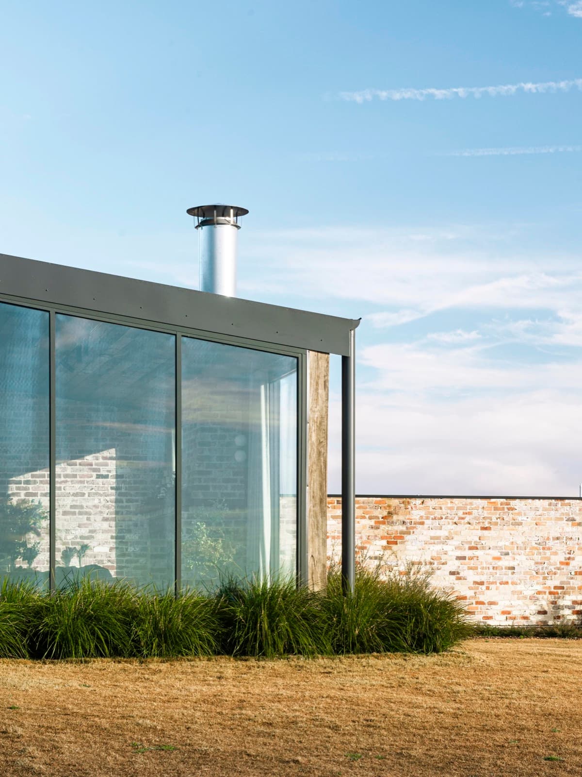 Glass corner of the Pavilion beside its recycled-brick wall, native grasses catching the morning sun