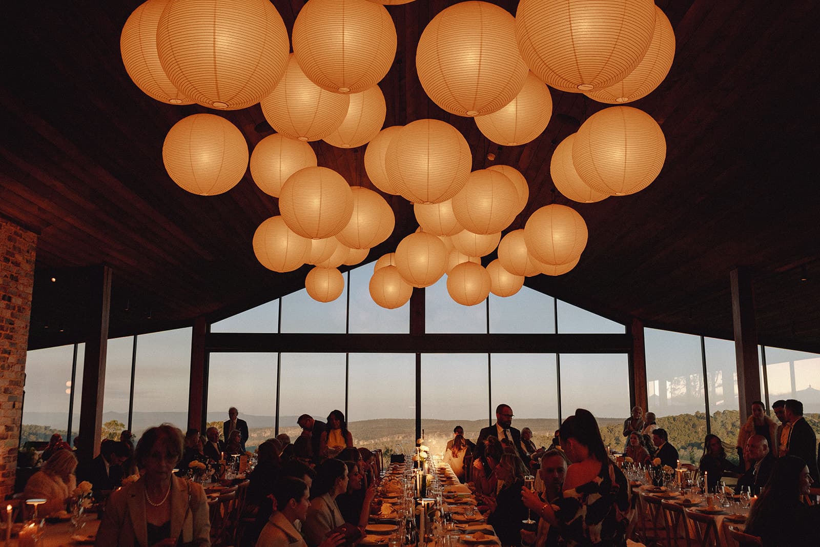 Paper lanterns glowing above the long table in the Pavilion, guests seated beneath the gabled glass with the escarpment beyond