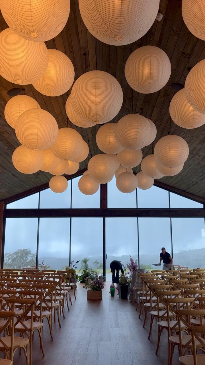 The Pavilion set for a wet-weather ceremony: cross-back chairs in rows either side of a centre aisle, paper lanterns overhead, mist against the glass gable