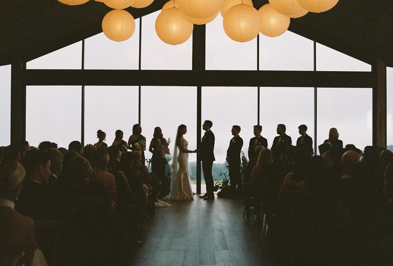 A wedding ceremony inside the glass Pavilion on a wet day, the couple silhouetted against the fog-filled gable window with guests seated on both sides of the aisle