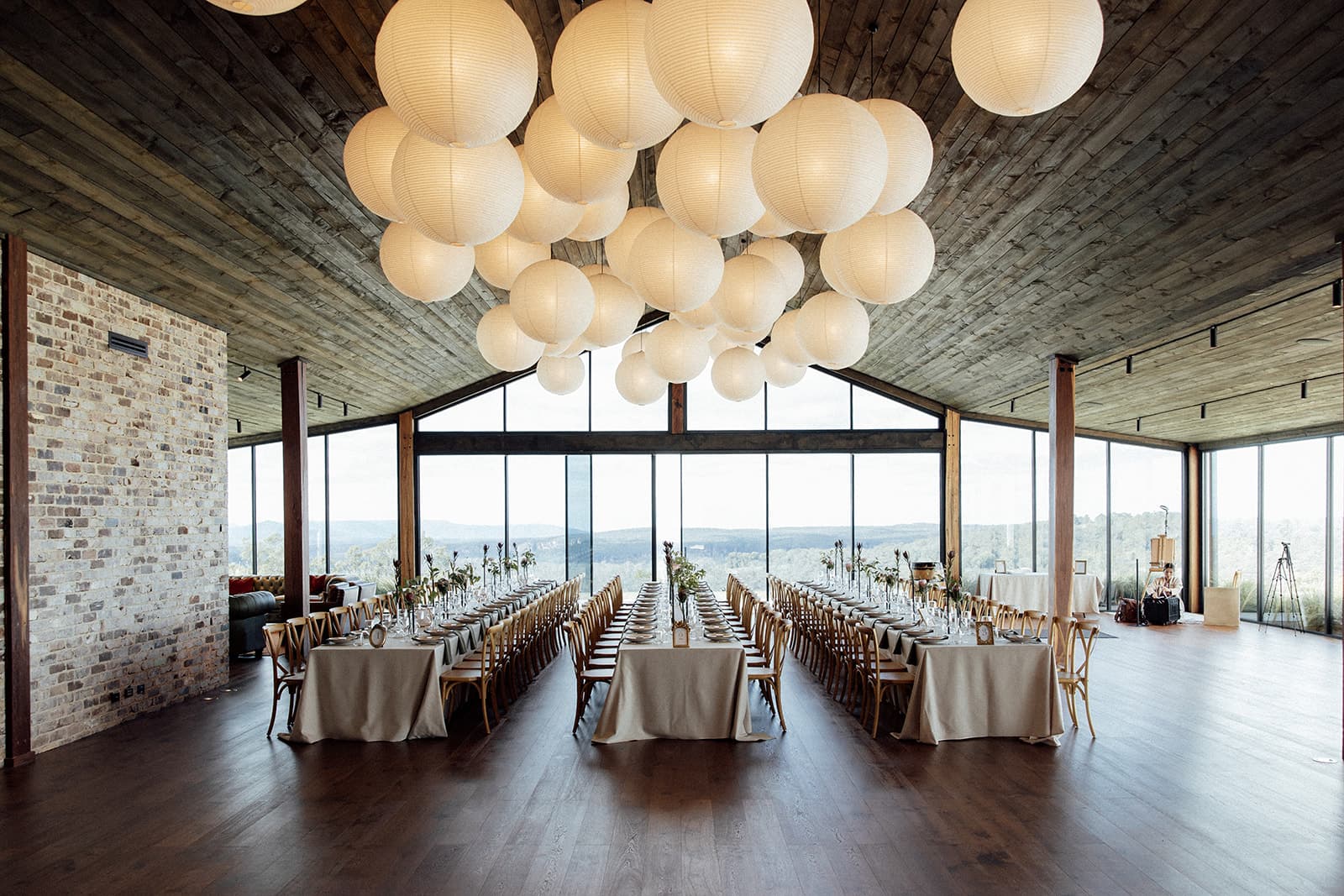 The Pavilion set for a wedding dinner: three long runs of tables with cross-back chairs, paper lanterns overhead, the timber posts standing clear and the escarpment through the glass gable