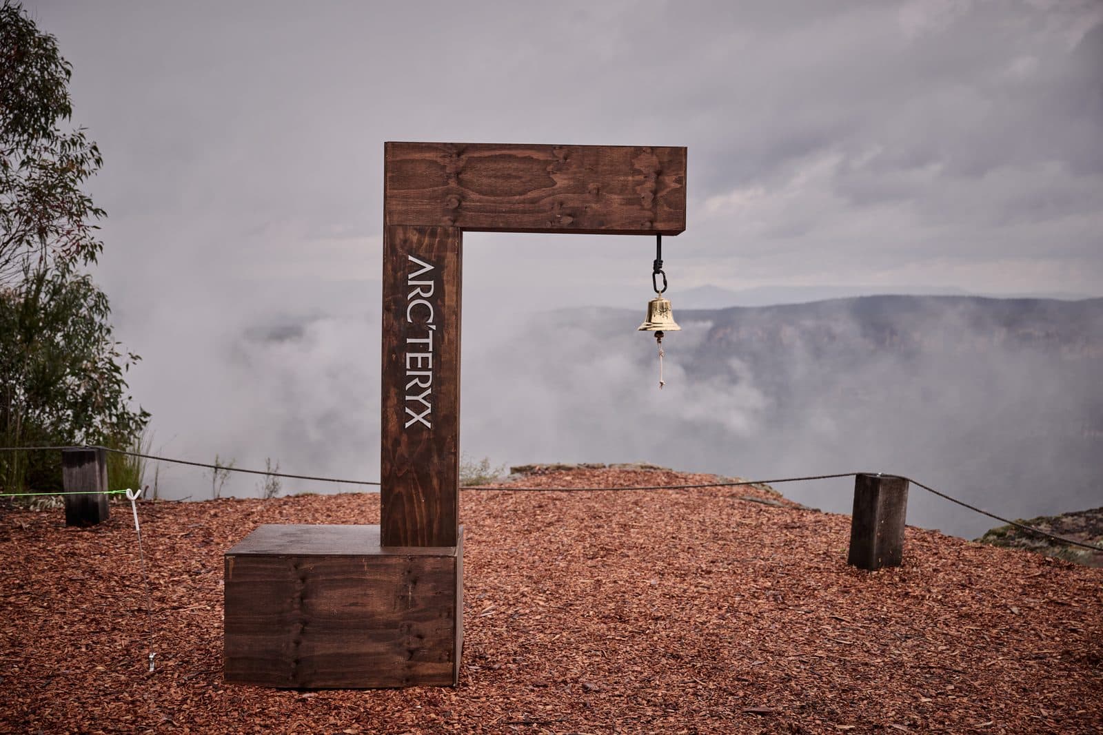 A timber post branded Arc'teryx at the escarpment edge, a brass bell hanging from it, cloud filling the gorge