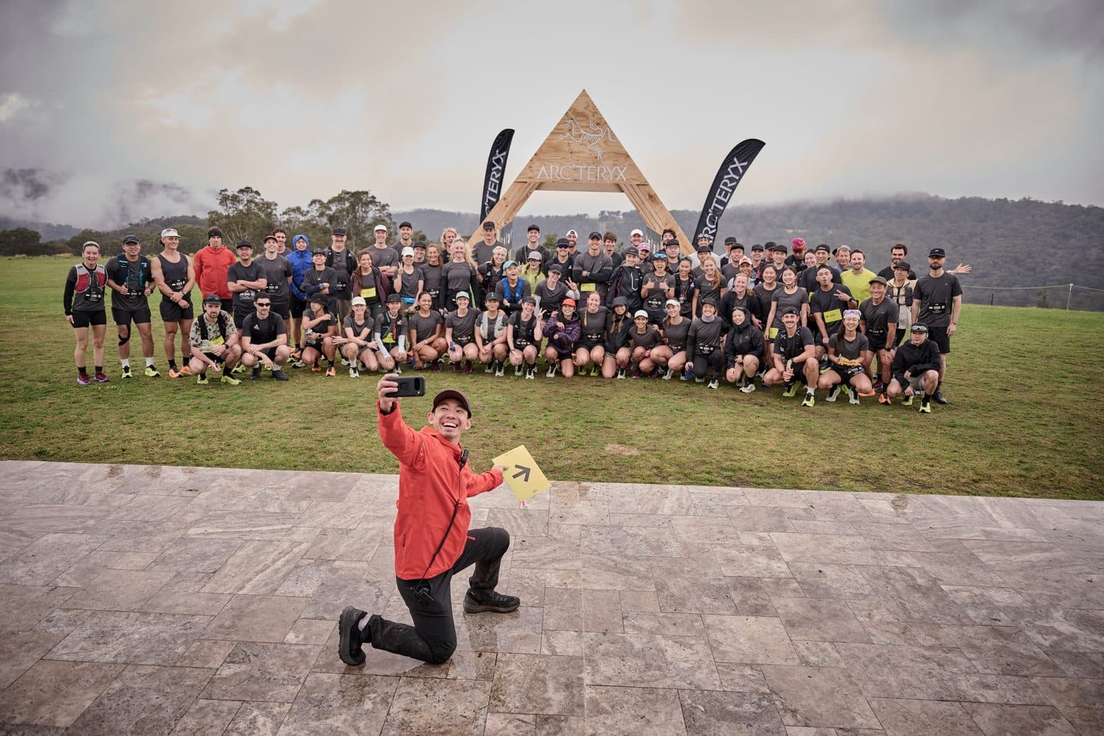 The whole field gathered under a timber A-frame arch on the lawn, the gorge in cloud behind