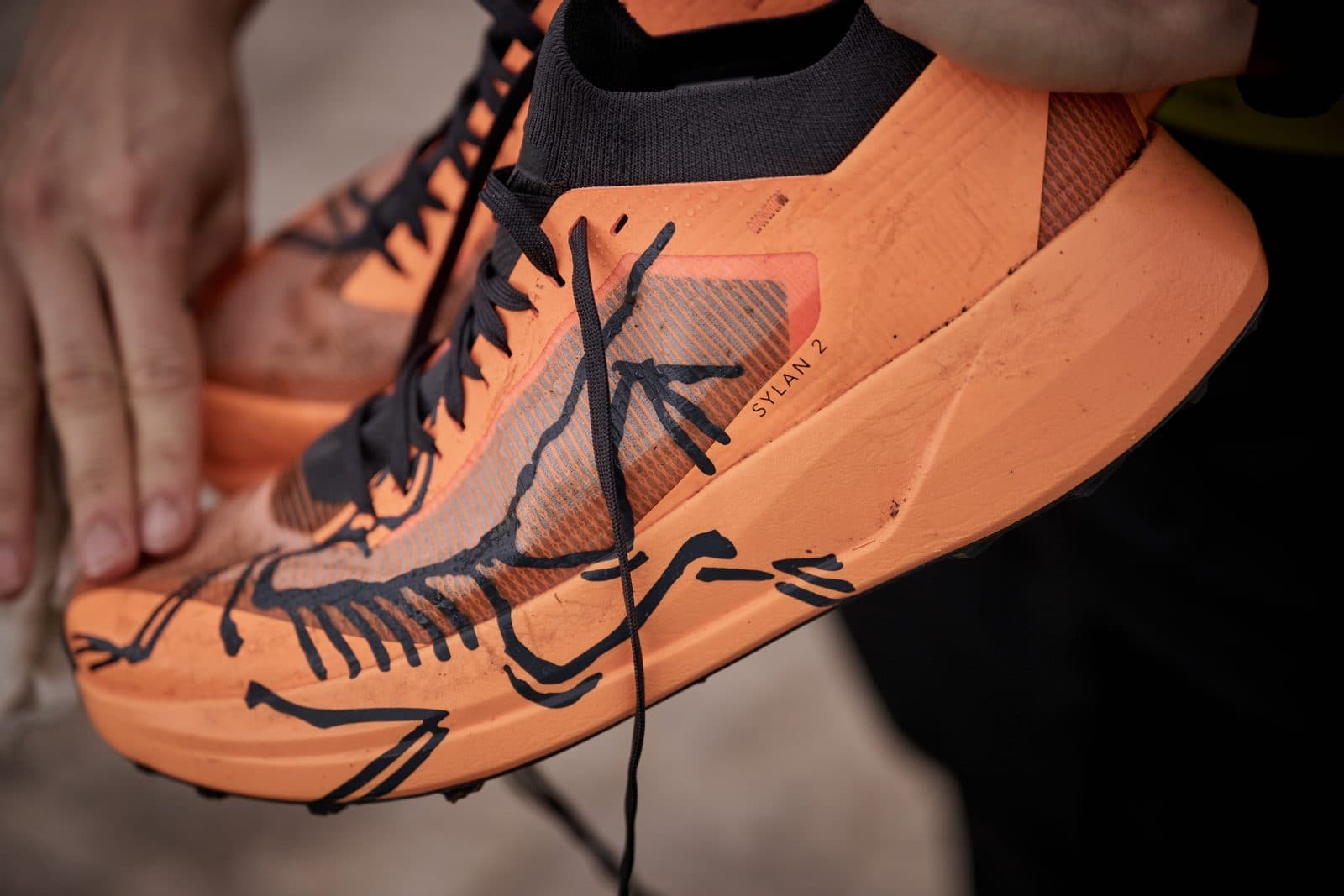 Hands holding an orange Sylan 2, the Arc'teryx bird drawn across its upper