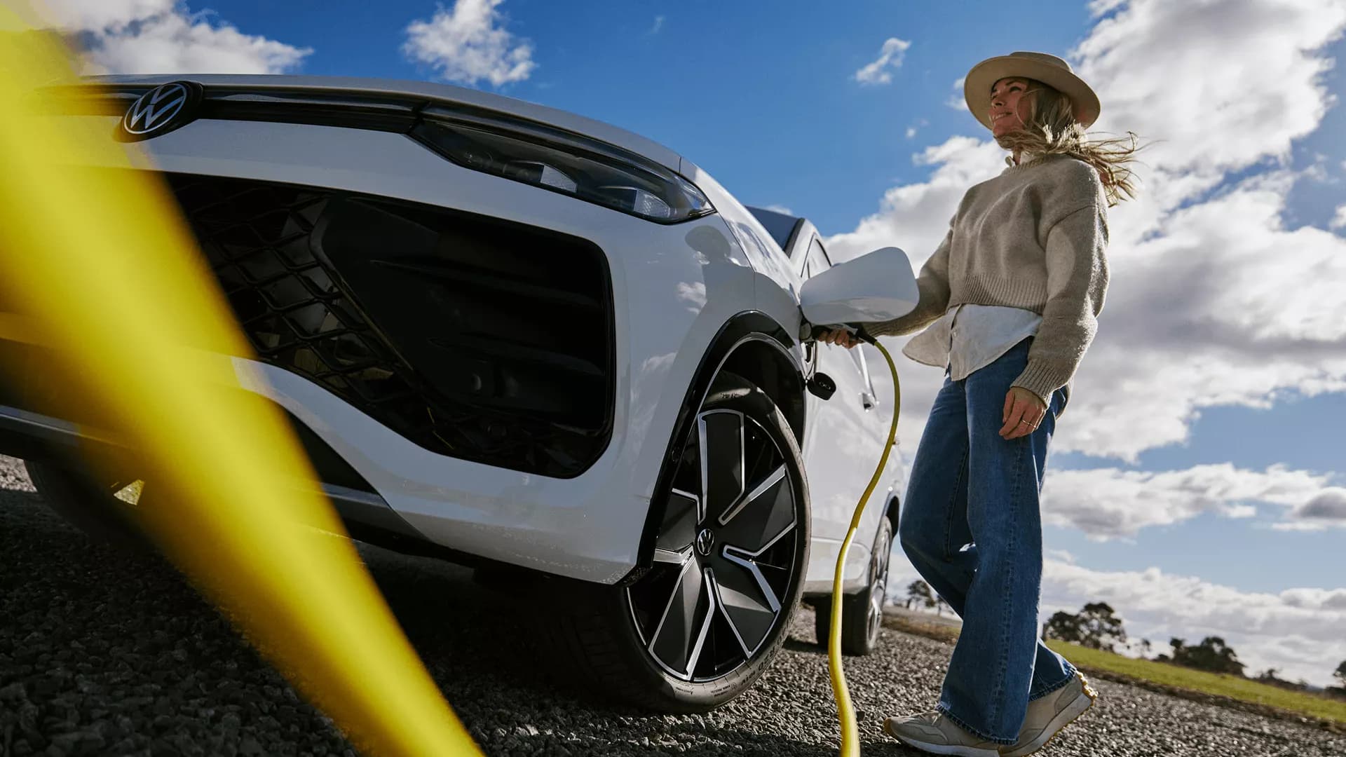 A woman plugging a charging cable into a white Volkswagen Tayron eHybrid under an open sky