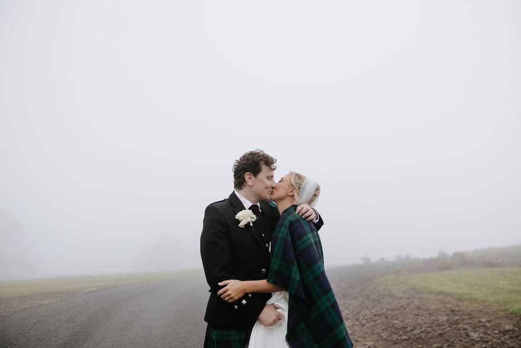 Anna and Calum kissing on the farm road in thick fog, his tartan over her shoulders