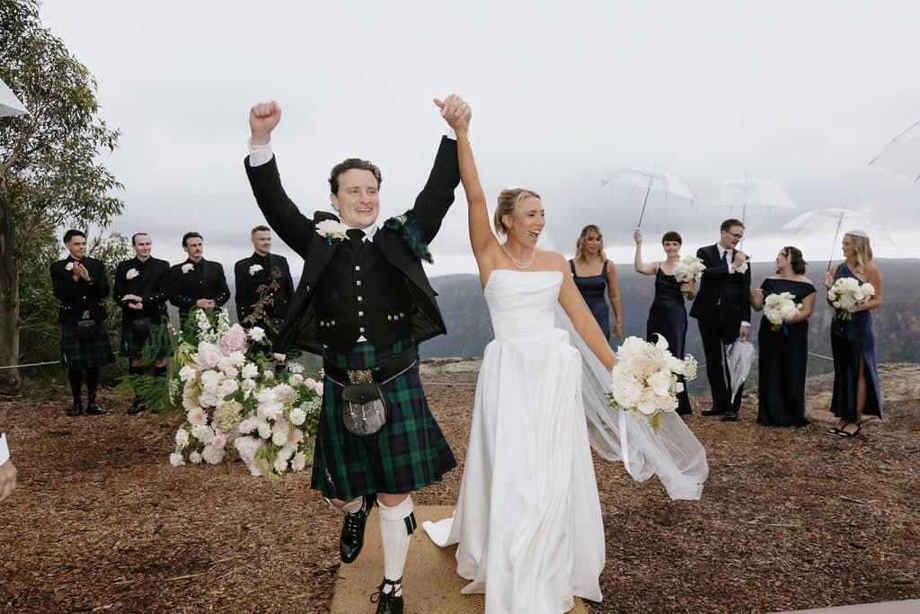 Anna and Calum with their arms raised as they come back up the aisle, the wedding party cheering behind them at The Cliffs