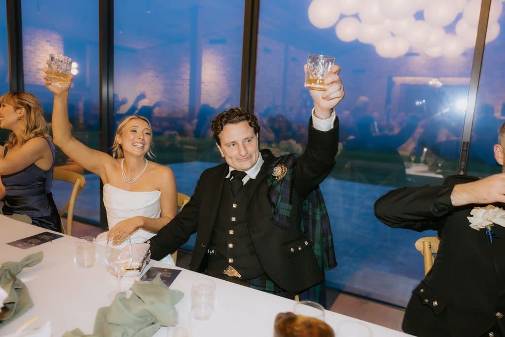 Anna and Calum raising their glasses at the table during speeches, dusk blue through the glass behind them