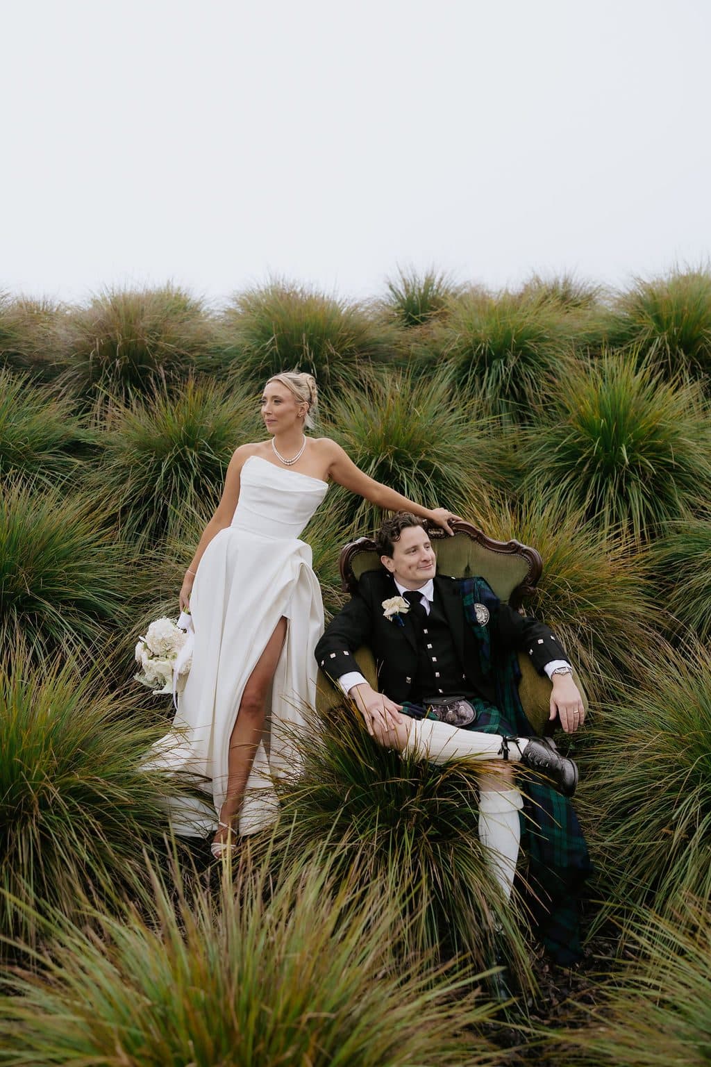 Calum in an armchair set among the tussock grasses, Anna standing beside him