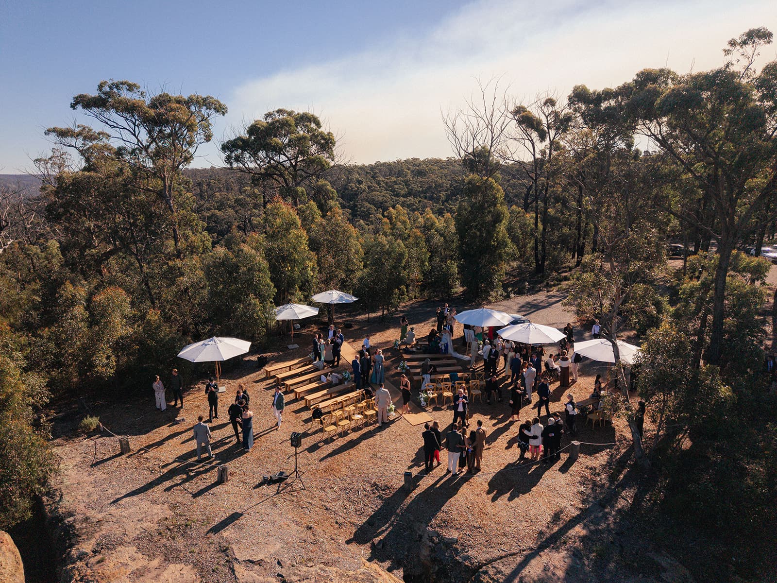 The clifftop ceremony site from above, guests gathering beneath white umbrellas among the gums