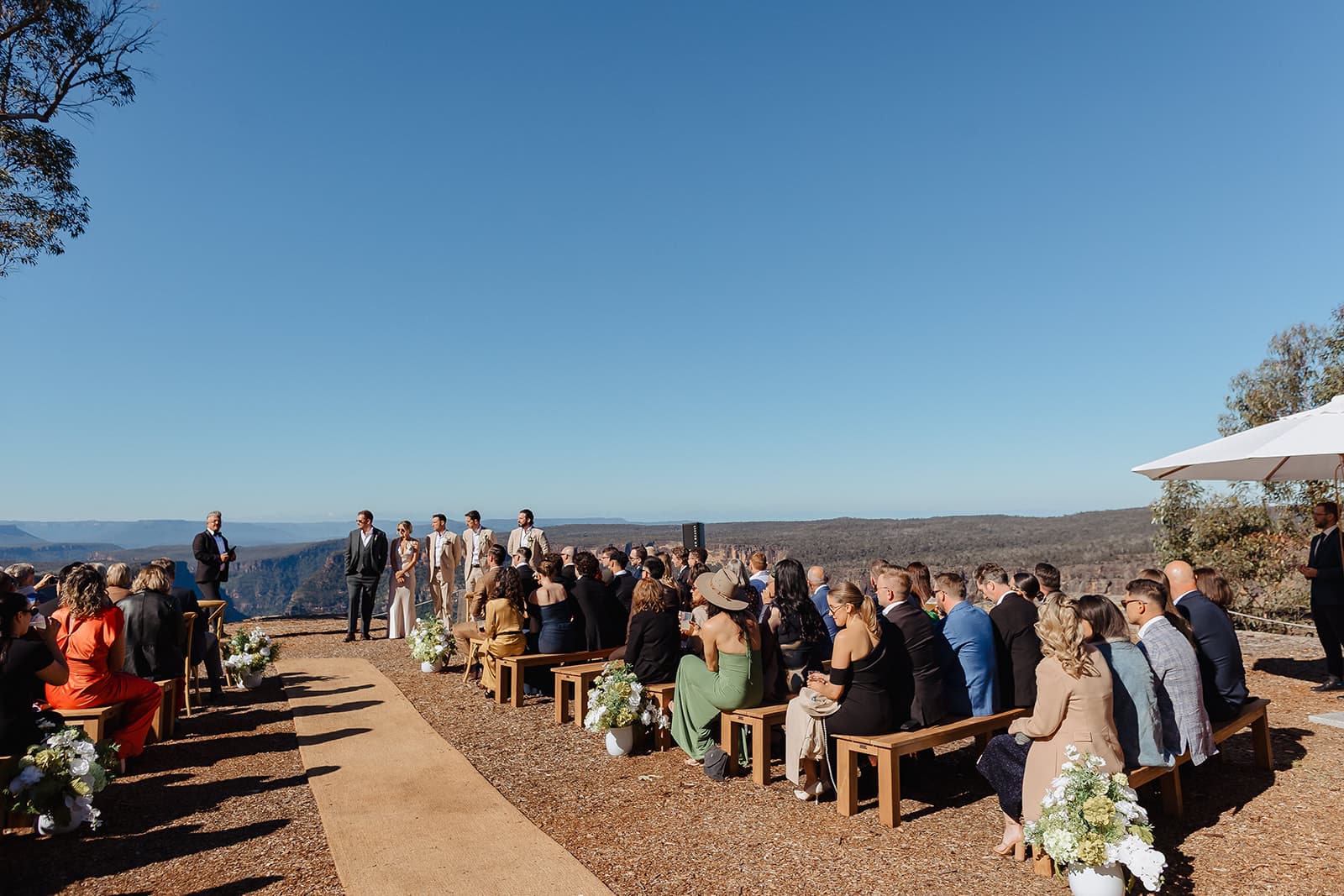 Guests seated on timber benches at the clifftop ceremony, the Shoalhaven gorge and escarpment stretching away behind the couple