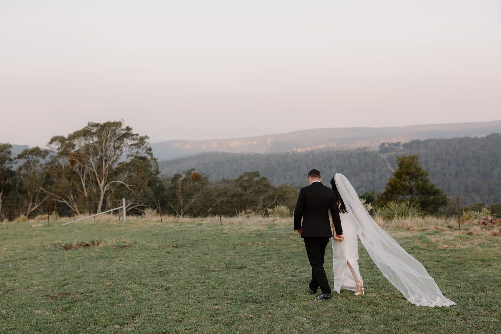 Jamae and Ben walking out across the paddock at dusk, her veil streaming behind her