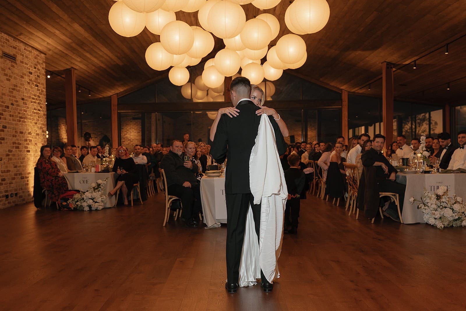 A couple dancing alone on the Pavilion floor under the lanterns, their guests watching from the long tables