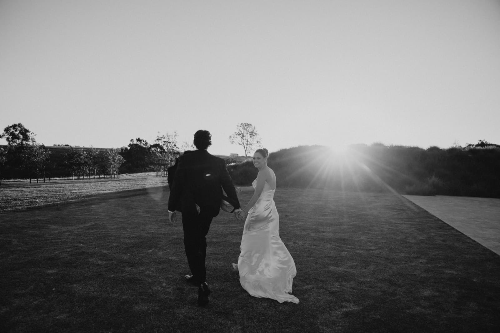 A couple walking hand in hand across the lawn toward the setting sun, the bride glancing back, in black and white