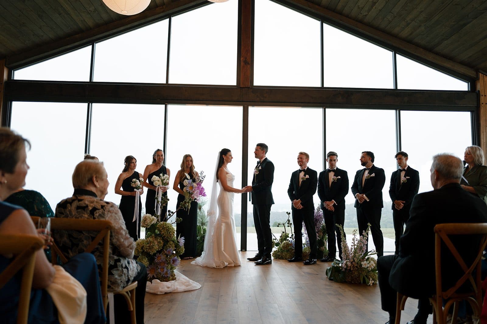 The ceremony from the back of the room, guests seated either side and the whole gable of glass behind the couple, full of fog