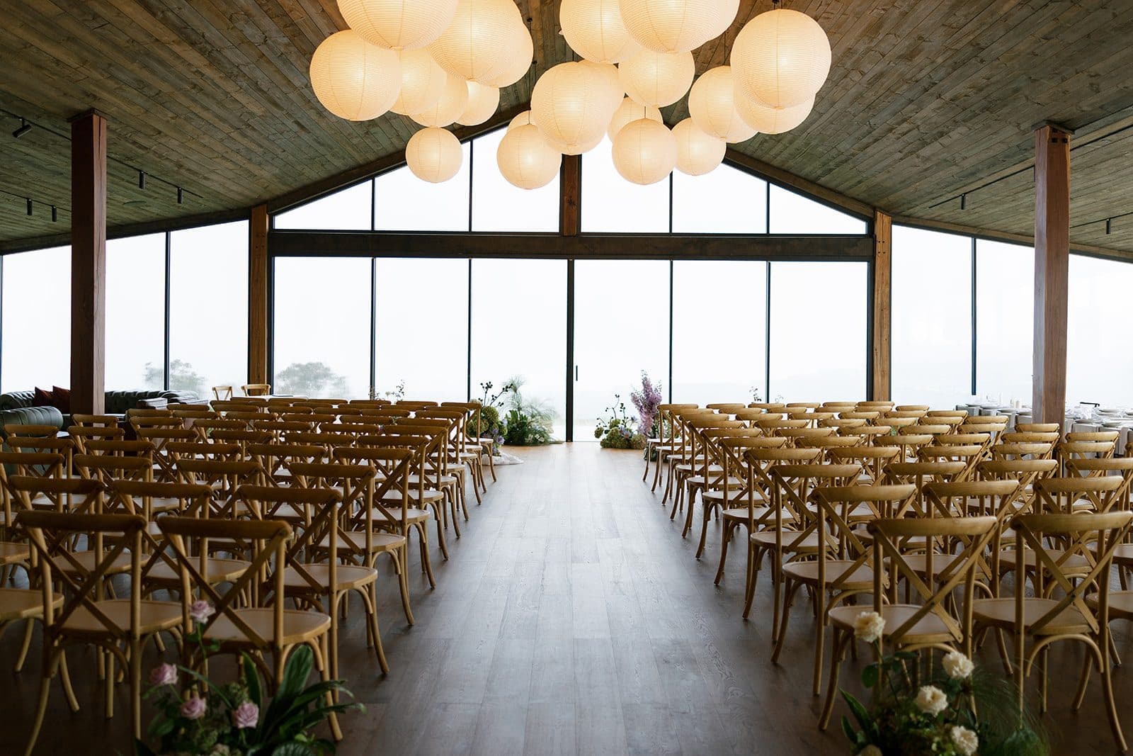 The Pavilion set for the ceremony, cross-back chairs in rows either side of a centre aisle running to the gable window, the lantern cluster lit above and cloud pressed against the glass