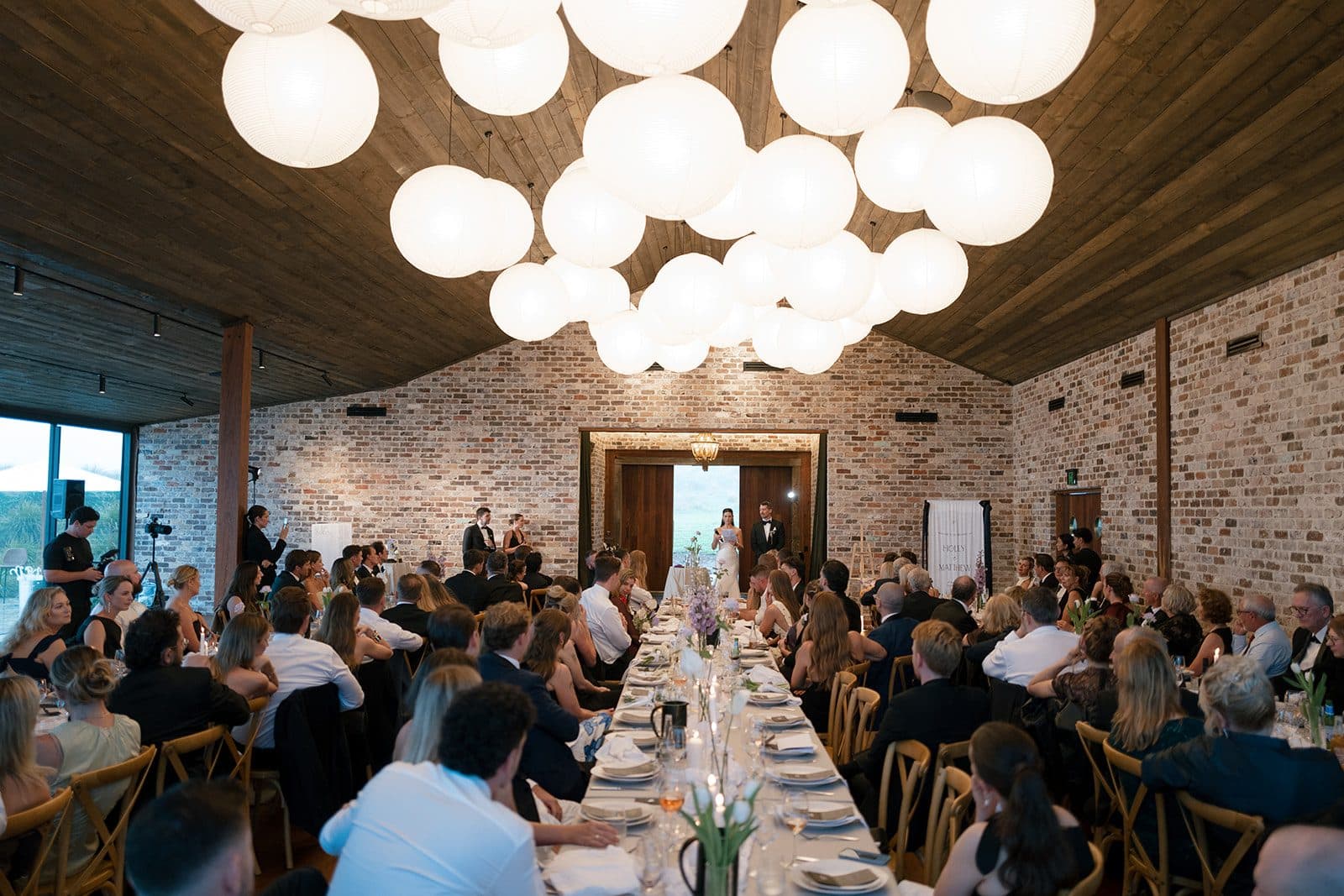 Dinner in The Pavilion, guests down both sides of the long tables under the lantern cluster, the timber doors open at the far end