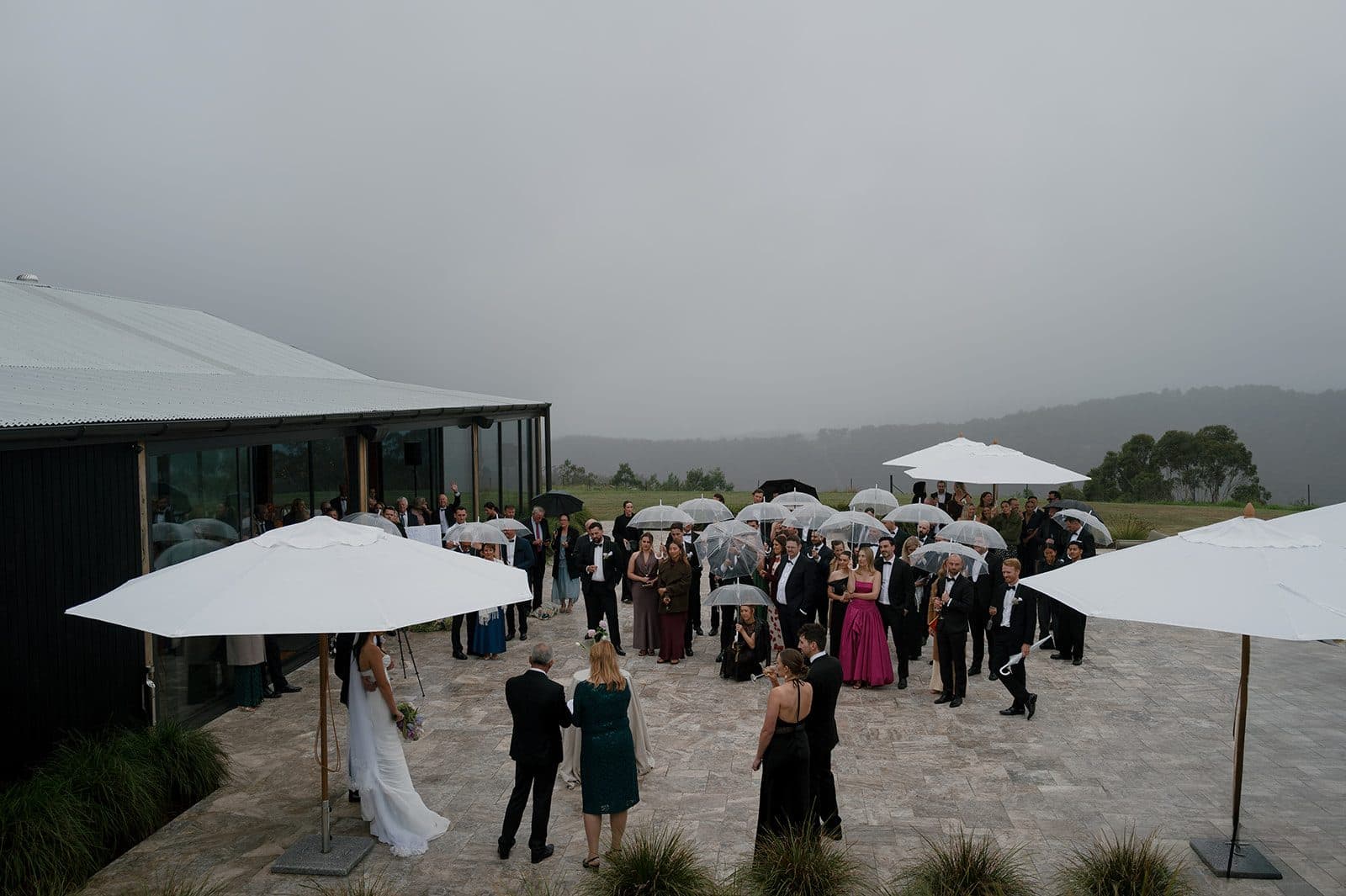 Cocktail hour on the terrace in the rain, guests under clear umbrellas and the Pavilion's white ones, the ridge behind them under low cloud