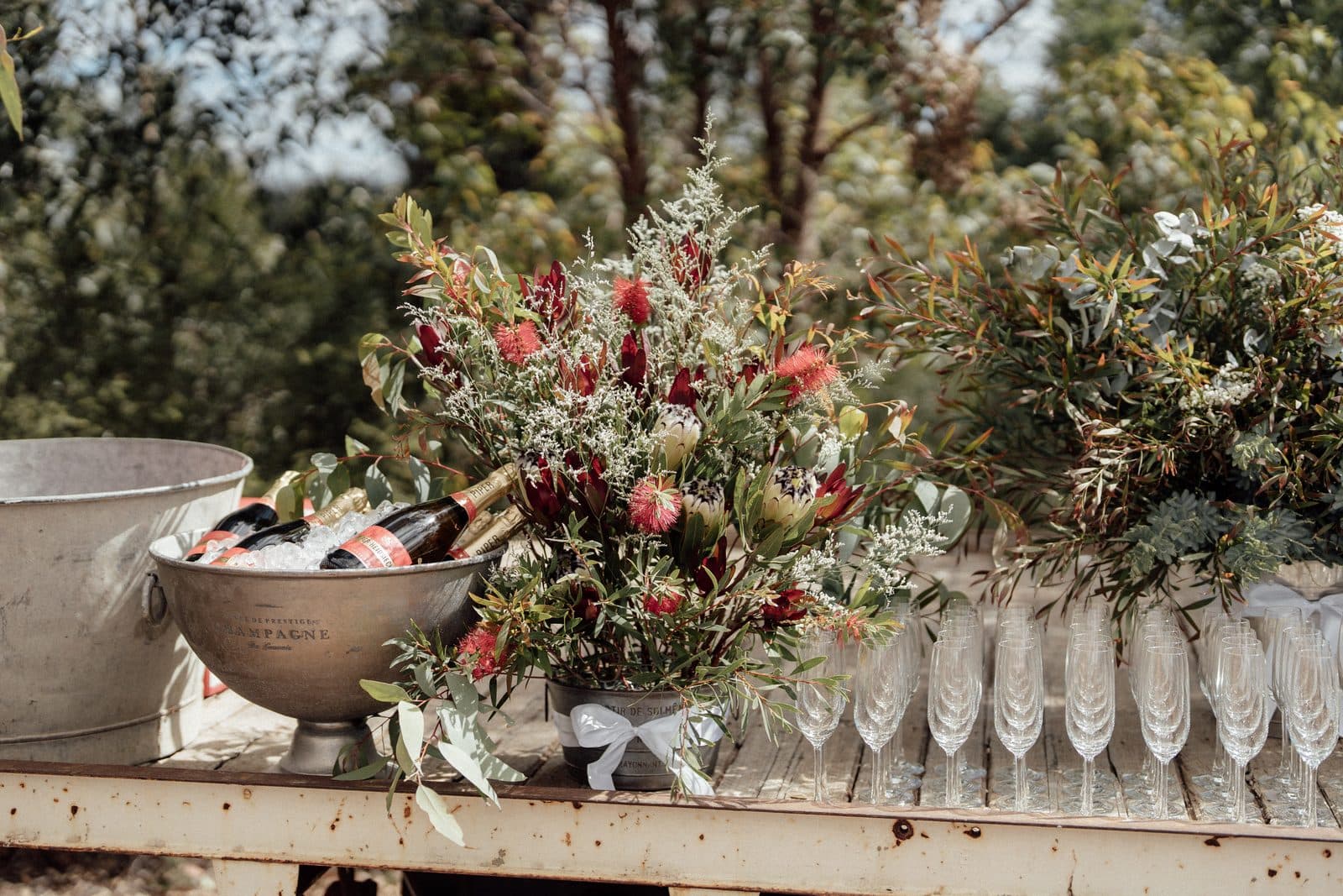 Champagne on ice in a silver bucket beside native florals and rows of flutes on an old timber flatbed